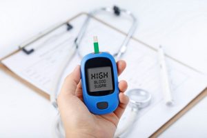 Hand Holding A Blood Glucose Meter Measuring Blood Sugar, The Background Is A Stethoscope And Chart File